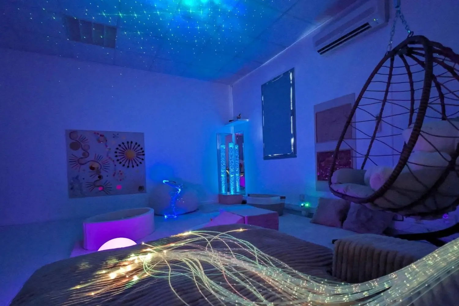 La salle Snoezelen de Sensoria plongée dans la pénombre : ciel étoilé projeté au plafond, fauteuil suspendu et fibres optiques lumineuses